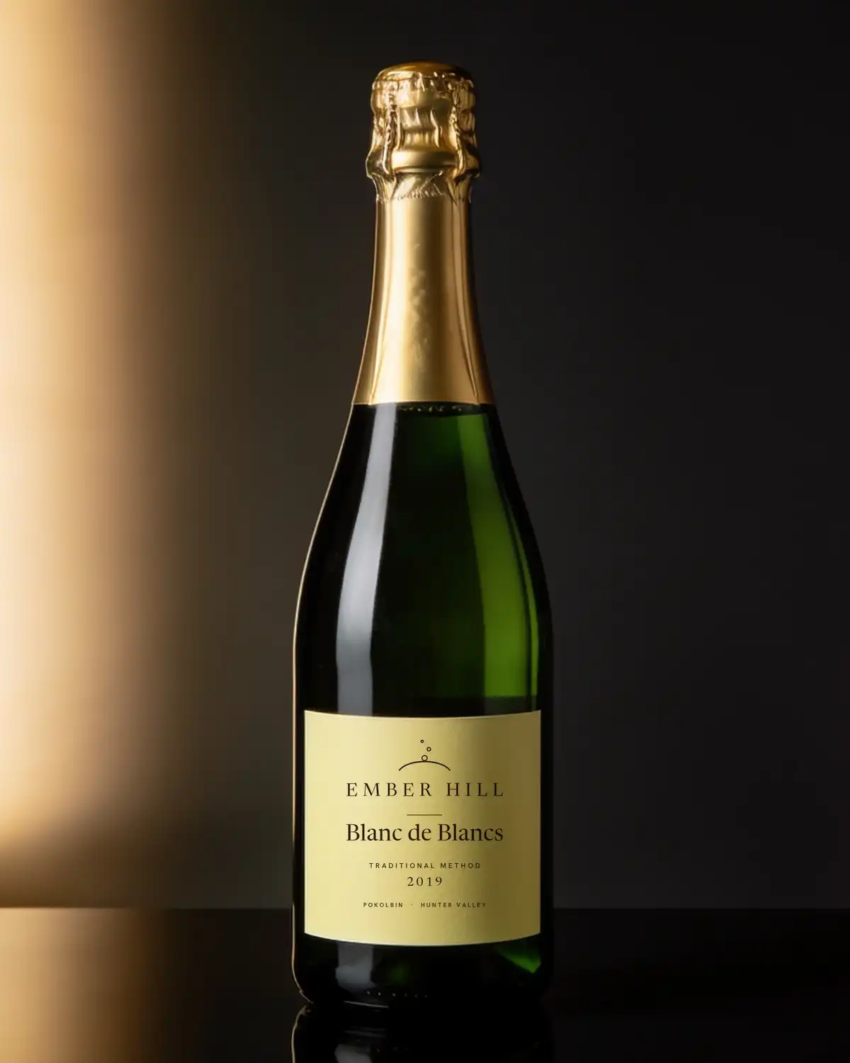 A bottle of Ember Hill Blanc de Blancs 2019, cream label, on a dark table